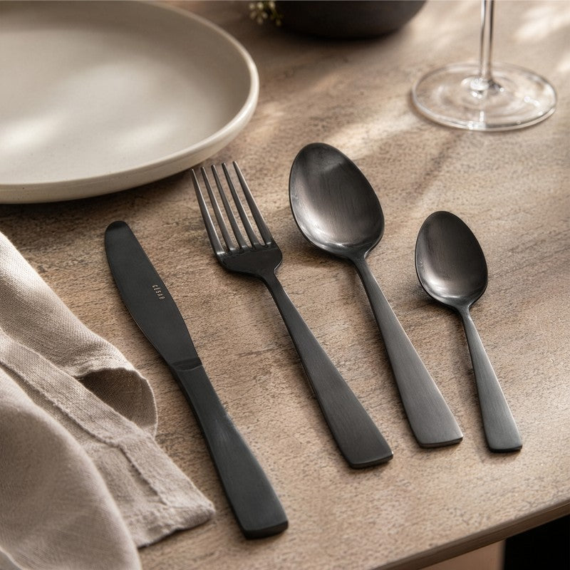 Mesa puesta con el set de cubiertos Lucia negro mate sobre superficie de madera, servilleta natural y copa de vino