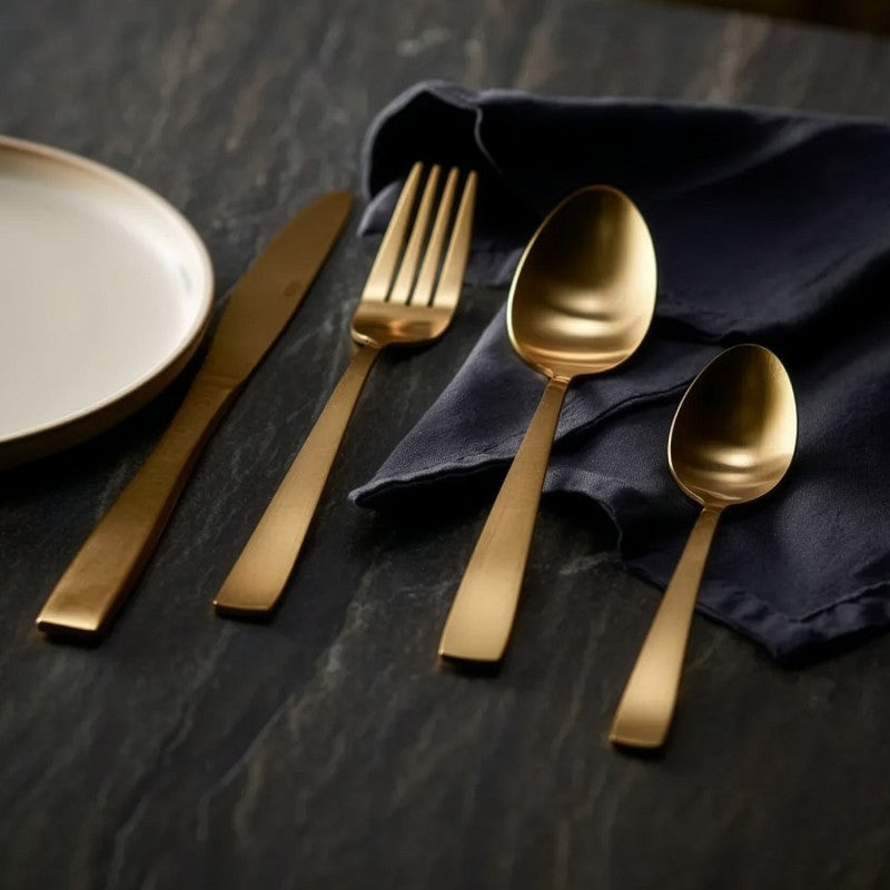 Mesa puesta con el set de cubiertos Lucia dorado mate sobre servilleta azul marino y superficie de madera oscura