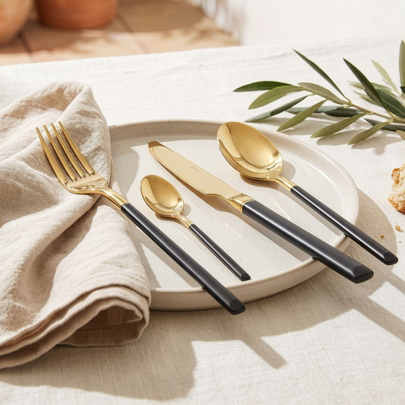 Mesa puesta con el set de cubiertos Noah dorado y negro sobre un plato de cerámica y servilleta de lino, ambientación elegante