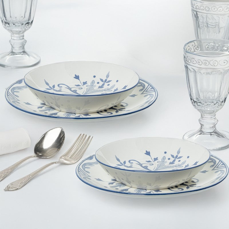 Plato y tazón de porcelana blanca con decoración floral azul dispuestos en mesa junto a copas y cubiertos