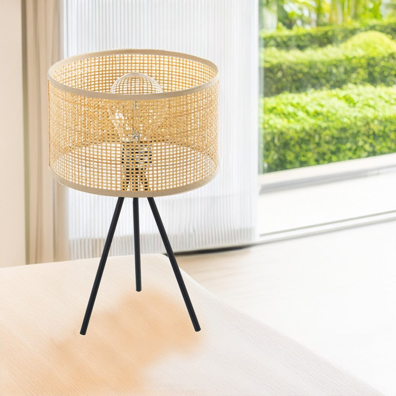 Lámpara de sobremesa de mimbre y metal sobre una mesa junto a un ventanal con vista a un jardín verde