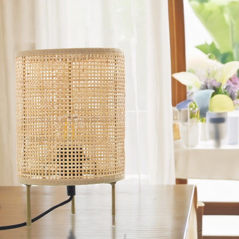 Lámpara de sobremesa con pantalla de rejilla de mimbre junto a una ventana con cortinas y flores