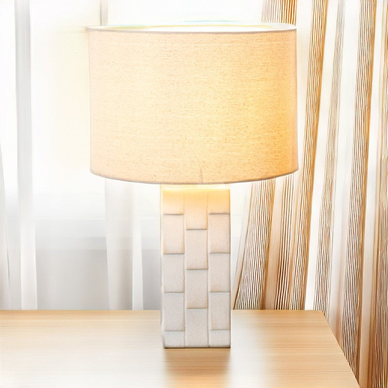 Lámpara de sobremesa cerámica blanca encendida sobre mesa de madera junto a una ventana con cortinas
