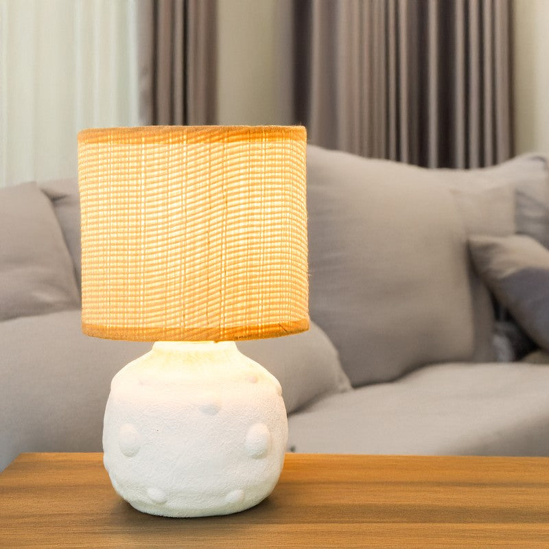 Lámpara de sobremesa cerámica blanca encendida sobre mesa de madera junto a un sofá con cojines, ambiente cálido
