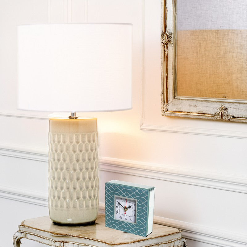 Lámpara de sobremesa cerámica crema encendida sobre mesilla en un dormitorio clásico junto a un reloj decorativo
